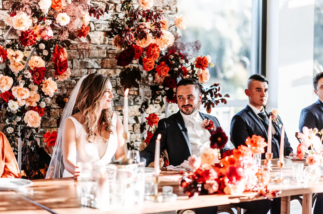 Bride and groom seated at flower-filled head table inside reception hall.