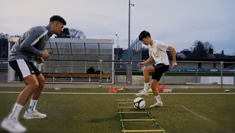 Fußballspieler macht eine Athletikübung mit einer Koordinationsleiter und wird von seinem Trainer angefeuert.
