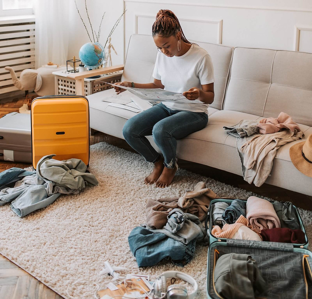 A person sitting on a sofa in a messy room with clothes and suitcases strewn about. The person is sitting amidst the mess on a sofa, engrossed in something they’re reading.