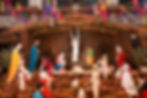 Nativity scene with colorful figures in various poses, set under a rustic shelter. Bright piñatas hang above; sheep and a manger are visible.