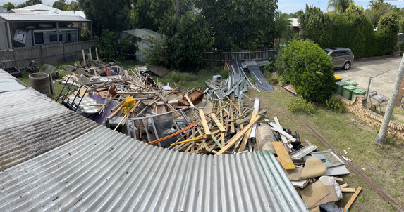 Aerial view from the roof of an old beach shack, showing the backyard cluttered with rubbish including wooden planks, carpet, windows, and furniture.