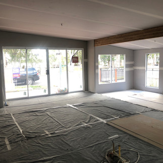 nside view of an open-plan house with a new layout. An exposed beam, new wall sheeting, large windows, and sliding doors are visible. Plastic sheeting covers the wooden floor for protection.