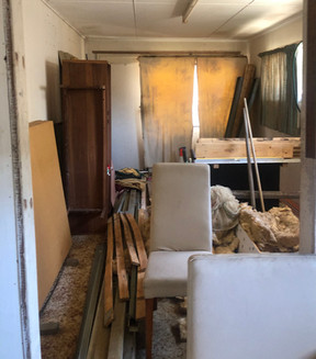 Interior of an abandoned house with old, dusty furniture, scattered pieces of wood, tattered curtains, stained carpet, and worn dining chairs. The room is cluttered with rubbish and debris.