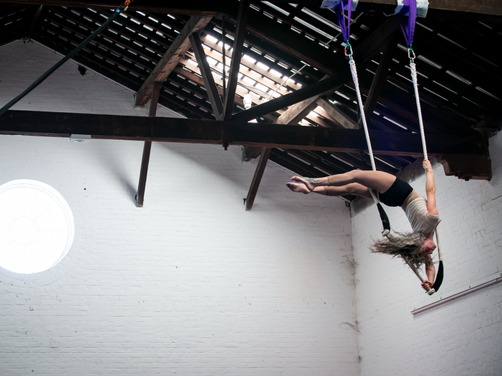 a circus performer doing a trick on a trapeze. She is in a white circus space with a round window and exposed beams.