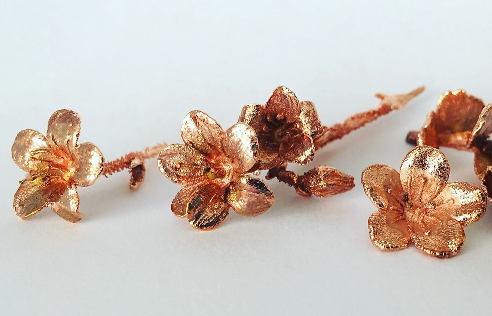 Three copper electroformed cherry blossom flowers on a branch against a white background, showing fine surface detail in bright copper.