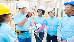 A group of individuals are at a construction site. They're wearing light blue shirts and are smiling. They appear to be volunteers at a construction build project.