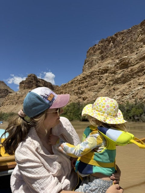An adult in a sun hat holding a toddler in a life jacket and wide-brimmed hat on a raft