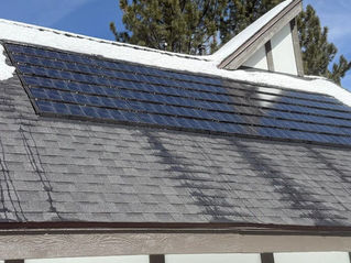 Picture of solar shingles atop a grey roof, with melting snow visible. Blue skies and a tall gree tree are visible in the background.