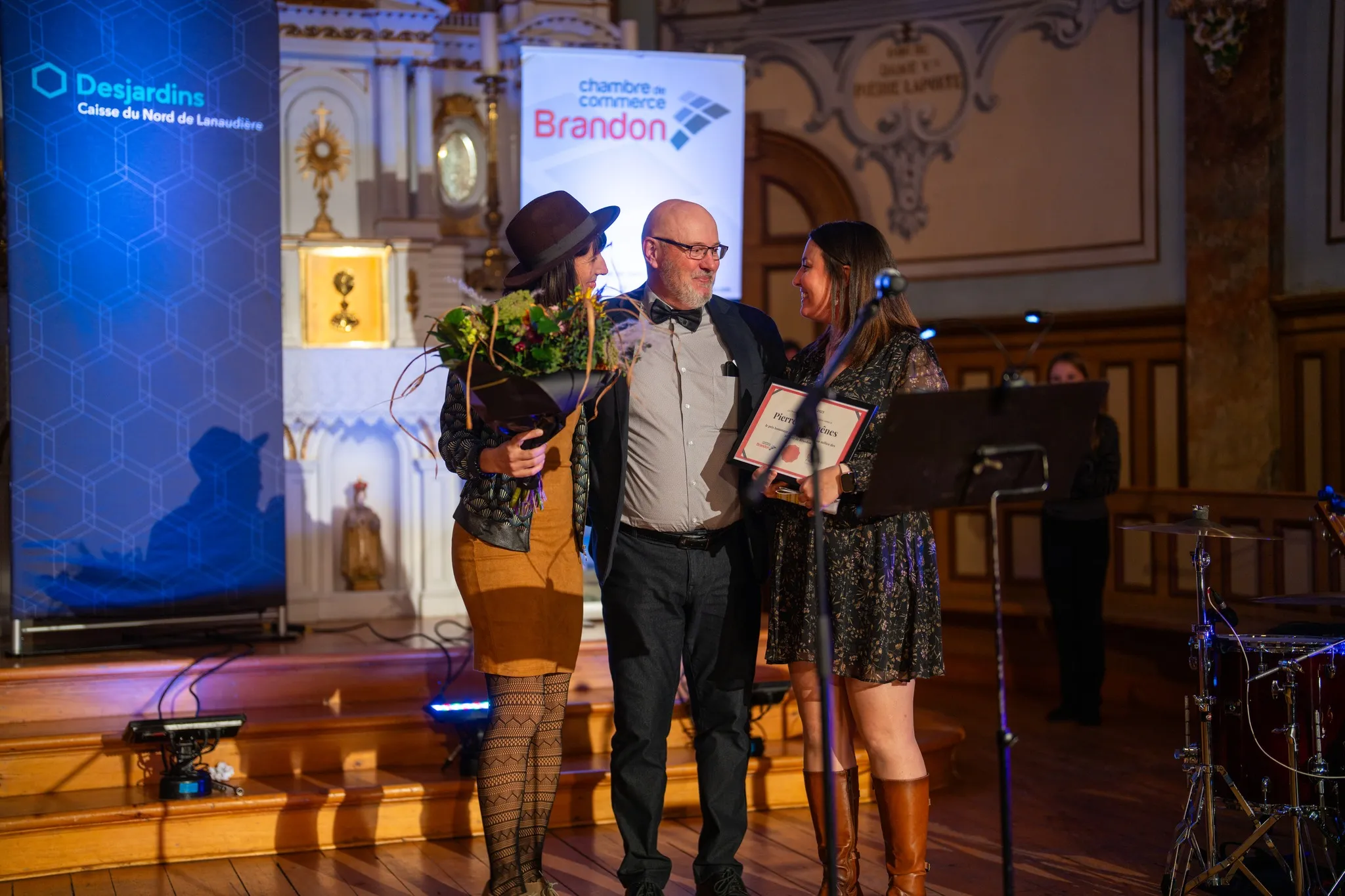Trois personnes réunies sur scène lors d’une remise de prix, avec un bouquet de fleurs et un certificat, devant le décor de l’événement organisé par la Chambre de commerce Brandon.