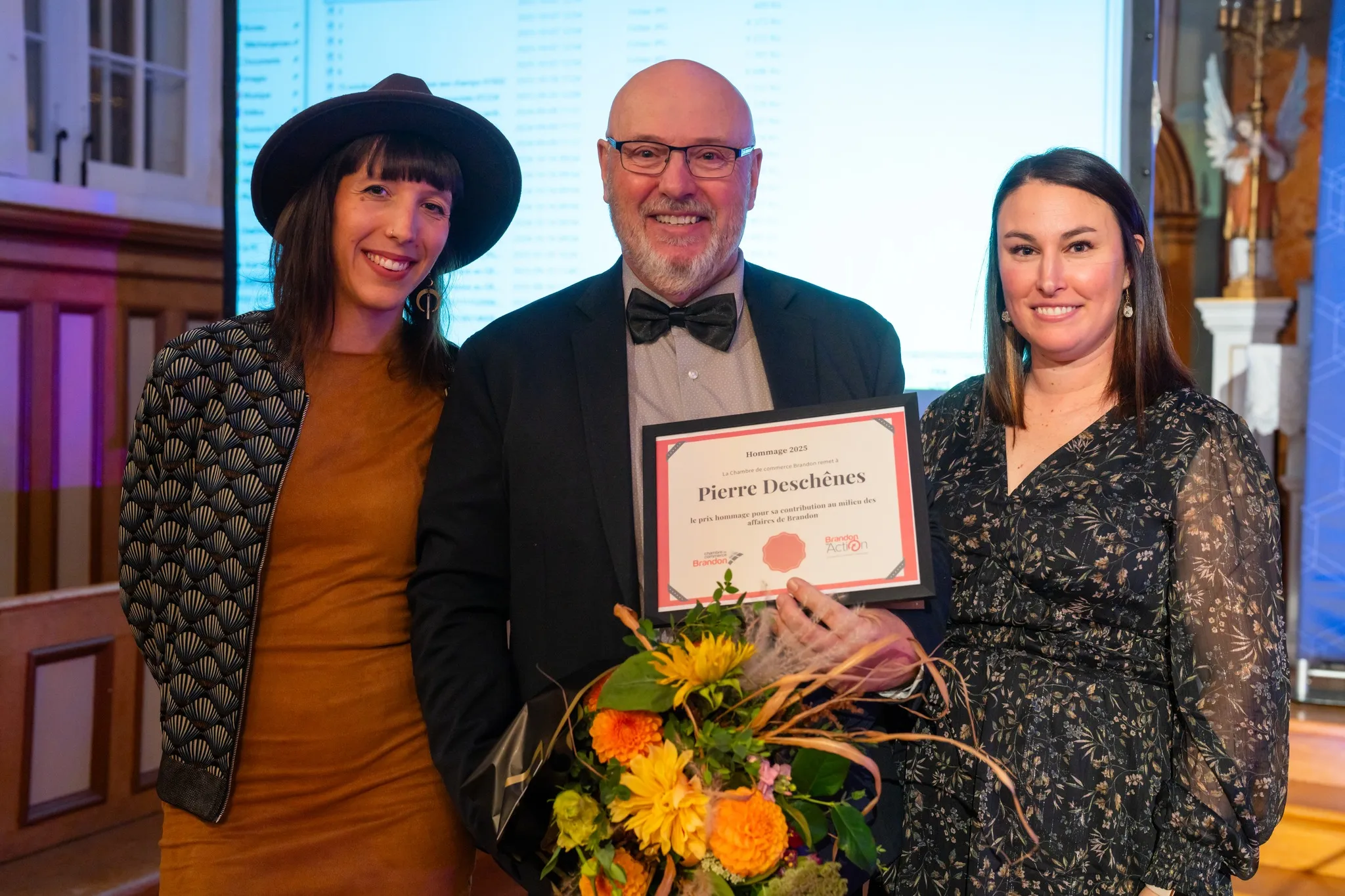 Trois personnes debout sur scène, souriantes, tenant un certificat et un bouquet de fleurs lors d’une remise de prix.