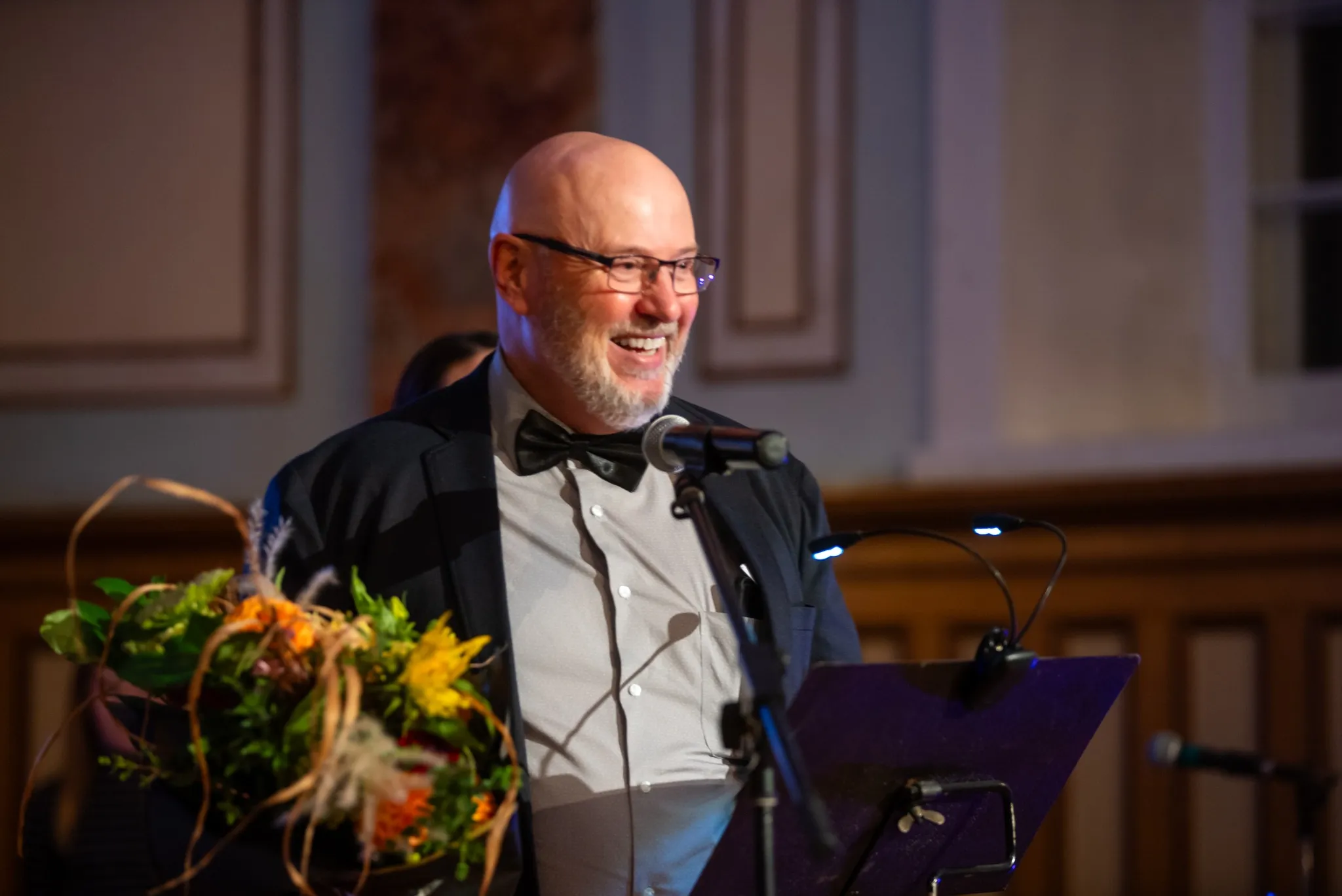 Un homme souriant en habit de gala prend la parole au micro, devant un pupitre décoré de fleurs, dans un lieu au décor patrimonial.