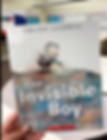 A hand holds the book "The Invisible Boy" by Trudy Ludwig. The cover shows a boy drawing. Classroom shelves are in the blurred background.