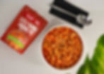 Five open cans of vegetables—peas, baked beans, chickpeas, corn, and mushrooms—on a wooden board with herbs and garlic on a white background.