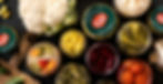 Various jars of pickled vegetables, including peppers, cucumbers, and beets, are arranged on a dark surface with a cauliflower.