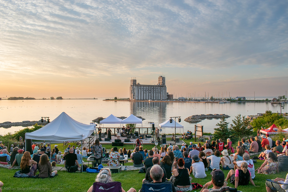 Outdoor concert crowd on a lakeside lawn at sunset, band on stage, with Collingwood Terminals silo across the water.