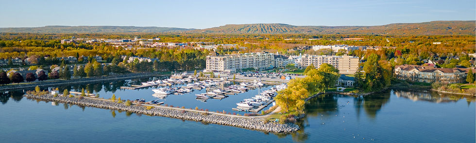 Aerial view of a calm marina with yachts, waterfront apartments, and autumn trees beside a lake and rolling hills.