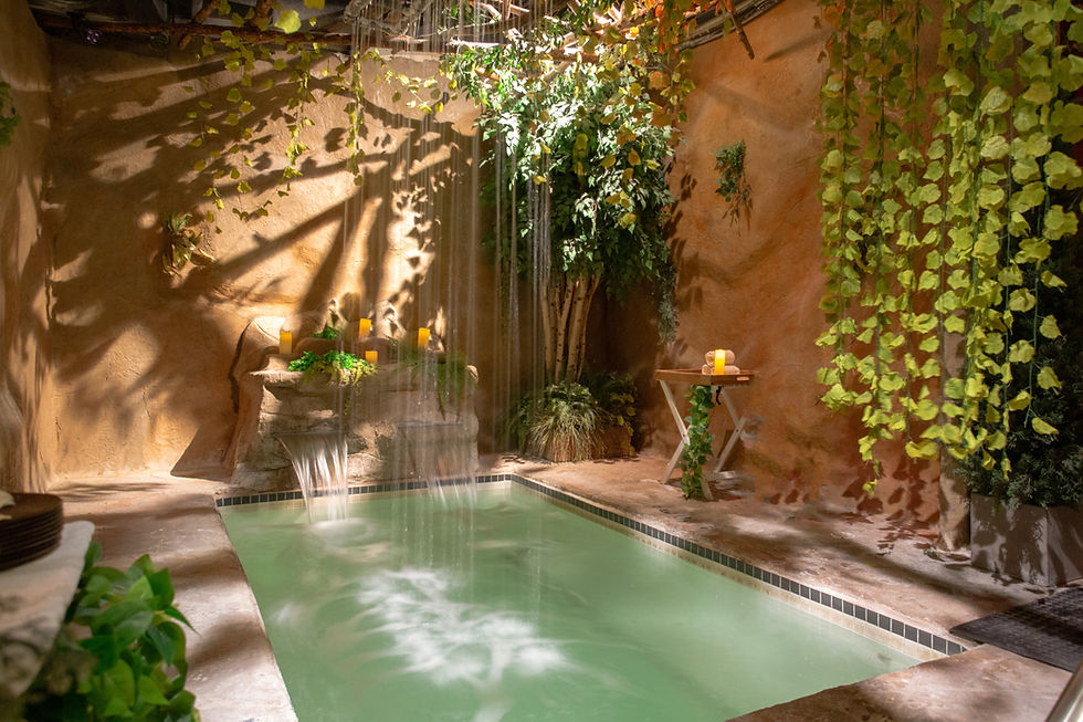 Indoor spa pool with gentle waterfalls, candles, and hanging vines, creating a warm, tranquil oasis with green water.