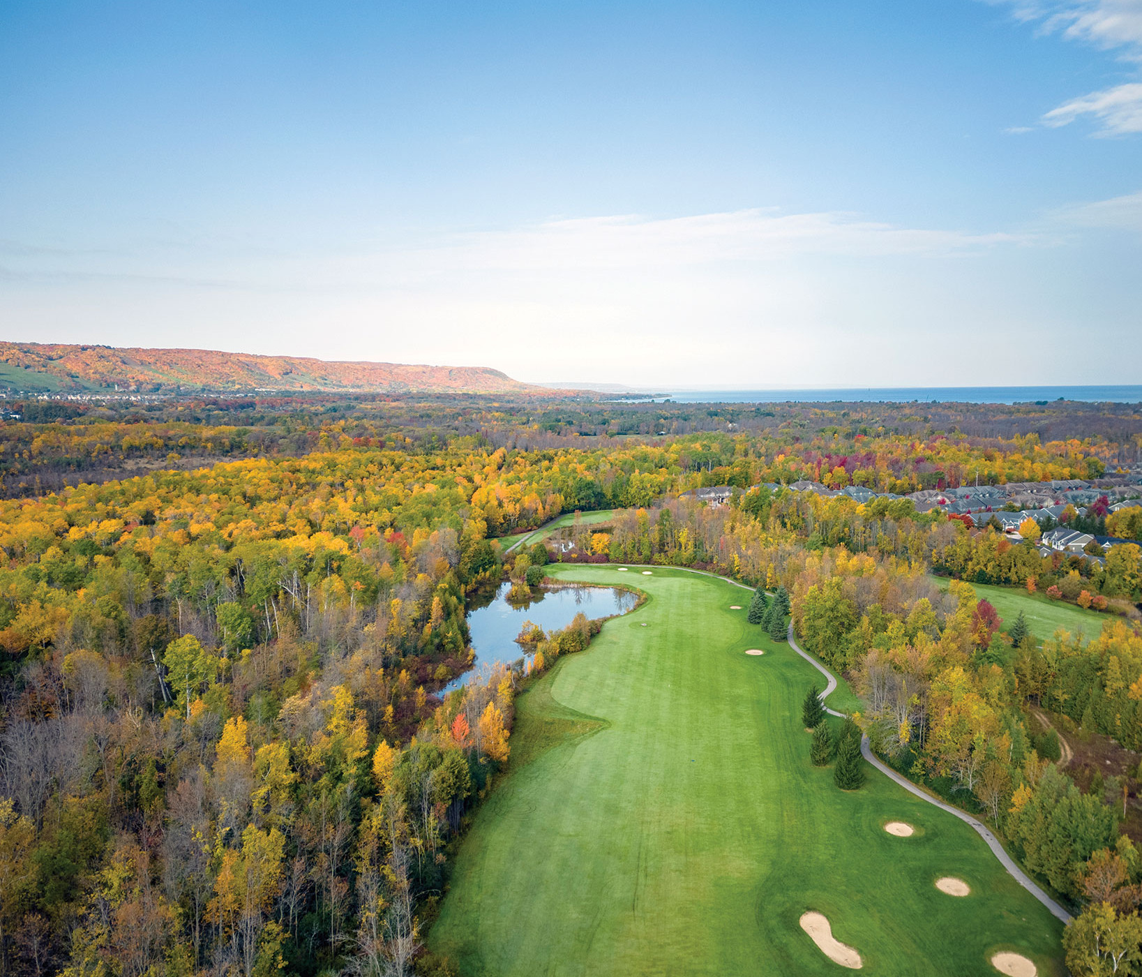 Aerial view of a golf course winding through autumn forest by a lake, with houses, distant hills, and blue sea under a clear sky