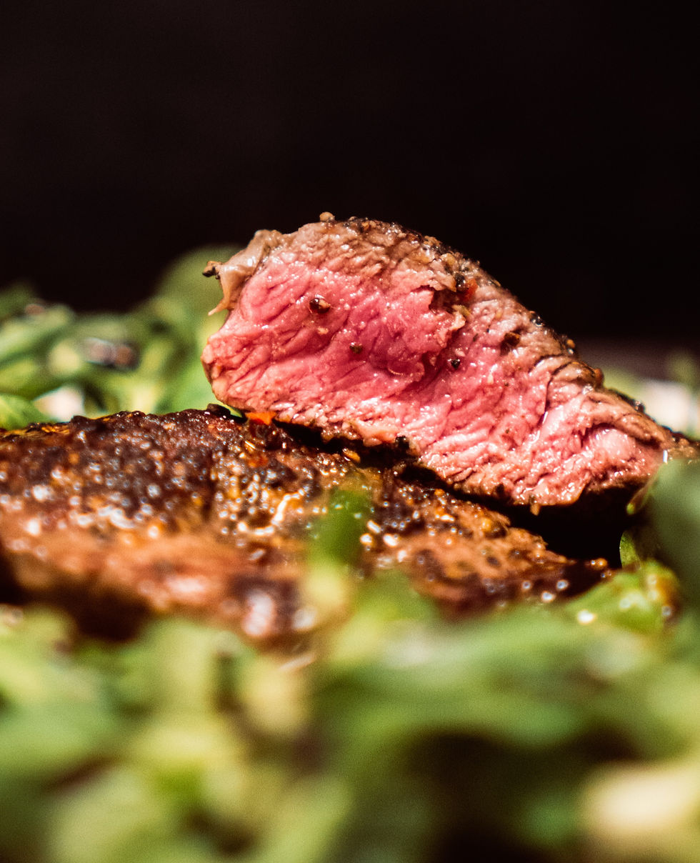 Close-up of a juicy, medium-rare steak slice atop greens on a dark background, showing a rich texture and appetizing color contrast.