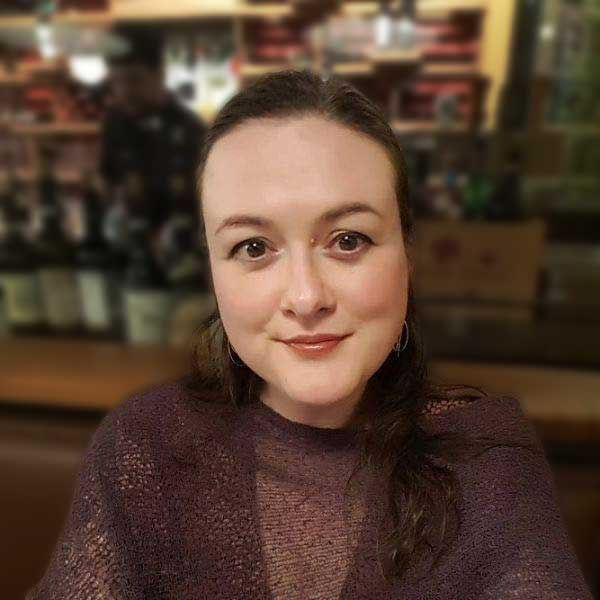 A woman wearing a purple top smiles at the camera in a cozy, warmly-lit room with shelves and blurred items in the background.