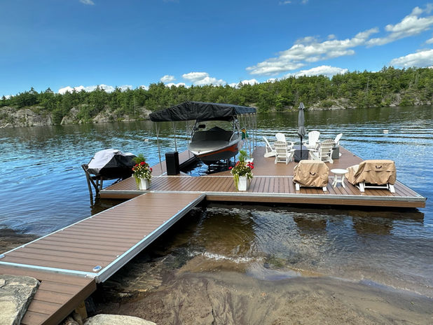 A custom pro series floating dock with a boat on a lift in the centre and furniture on the dock with pine trees in the background and a blue sky