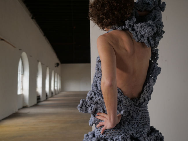 Robe crochetée aux doigts. Physionomie organique. Ruban jersey rayée blanc et bleu. Portée sur un corps, vu de dos. Un bras dénudé, l'autre bras avec une manche. Le dos est visible.