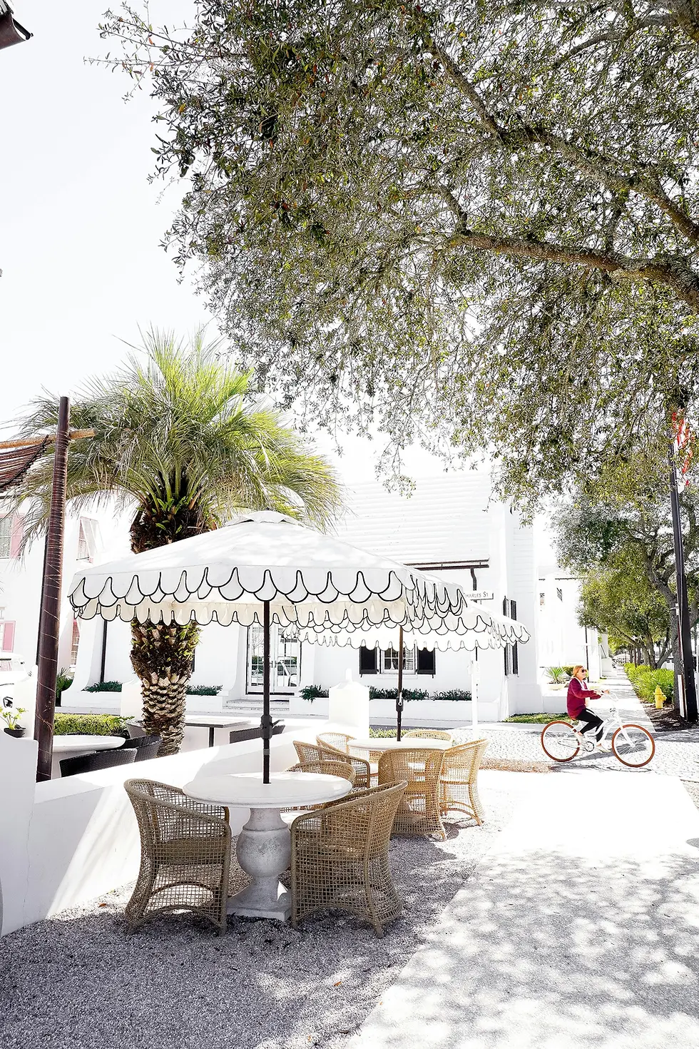 Wicker chairs and table under a scalloped umbrella, palm tree nearby, person in red biking on a sunny day, bright and serene ambiance.