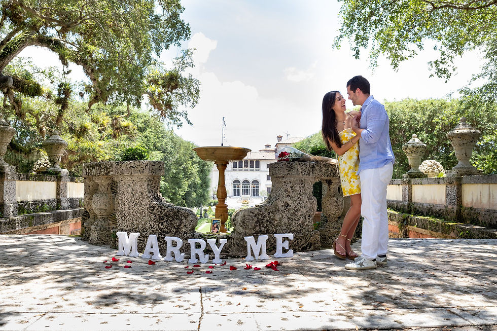 A couple embraces in a garden setting, surrounded by "Marry Me" letters and rose petals. They're near a stone fountain, exuding joy.