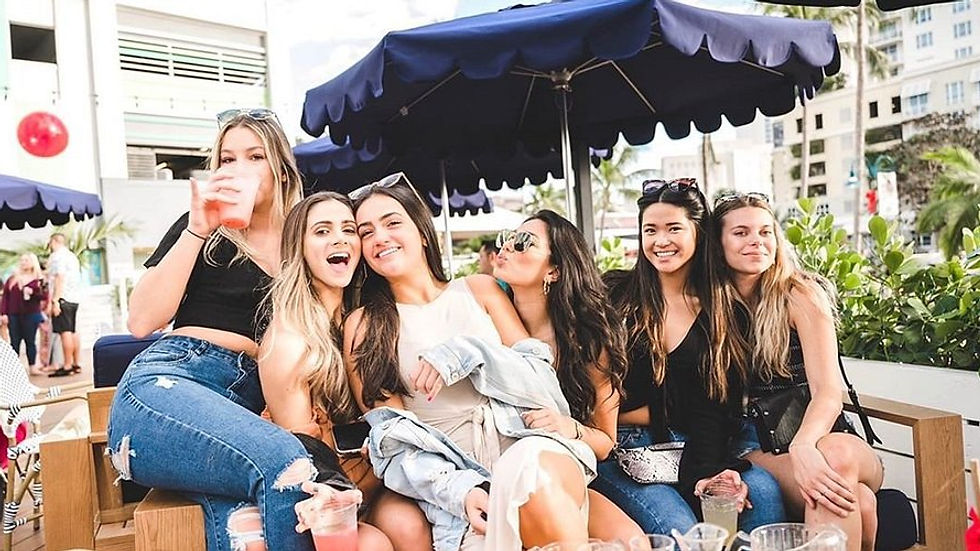 Six women sit together, smiling and holding drinks in an outdoor cafe with blue umbrellas. Casual attire, sunny day, lively mood.