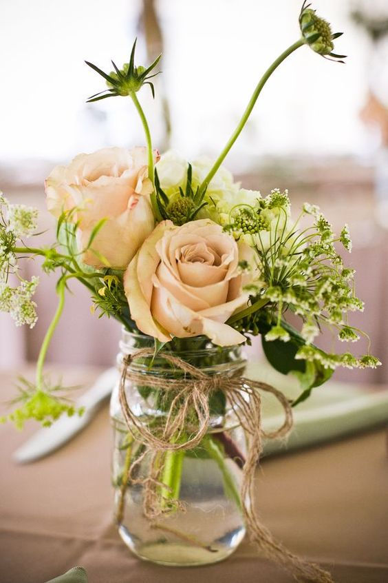 Pale pink roses in a glass jar tied with twine, surrounded by greenery. Soft, blurred background creates a serene, elegant atmosphere.