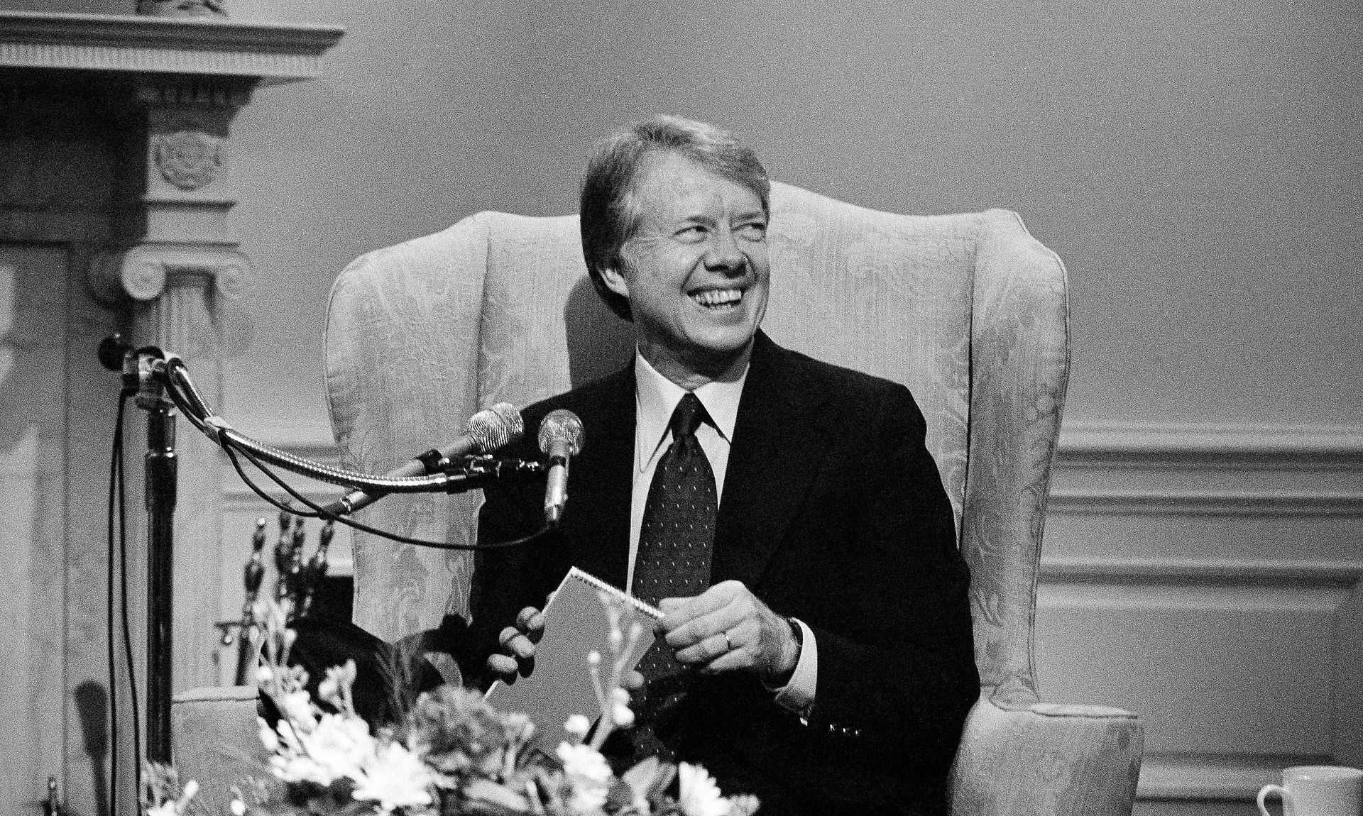 Jimmy Carter sitting in an armchair, wearing a suit and tie, smiling warmly in front of microphones, possibly during an interview or public appearance