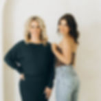 Two women smile in front of a light background. One wears a dark dress, the other a brown top with jeans. Relaxed, friendly mood.