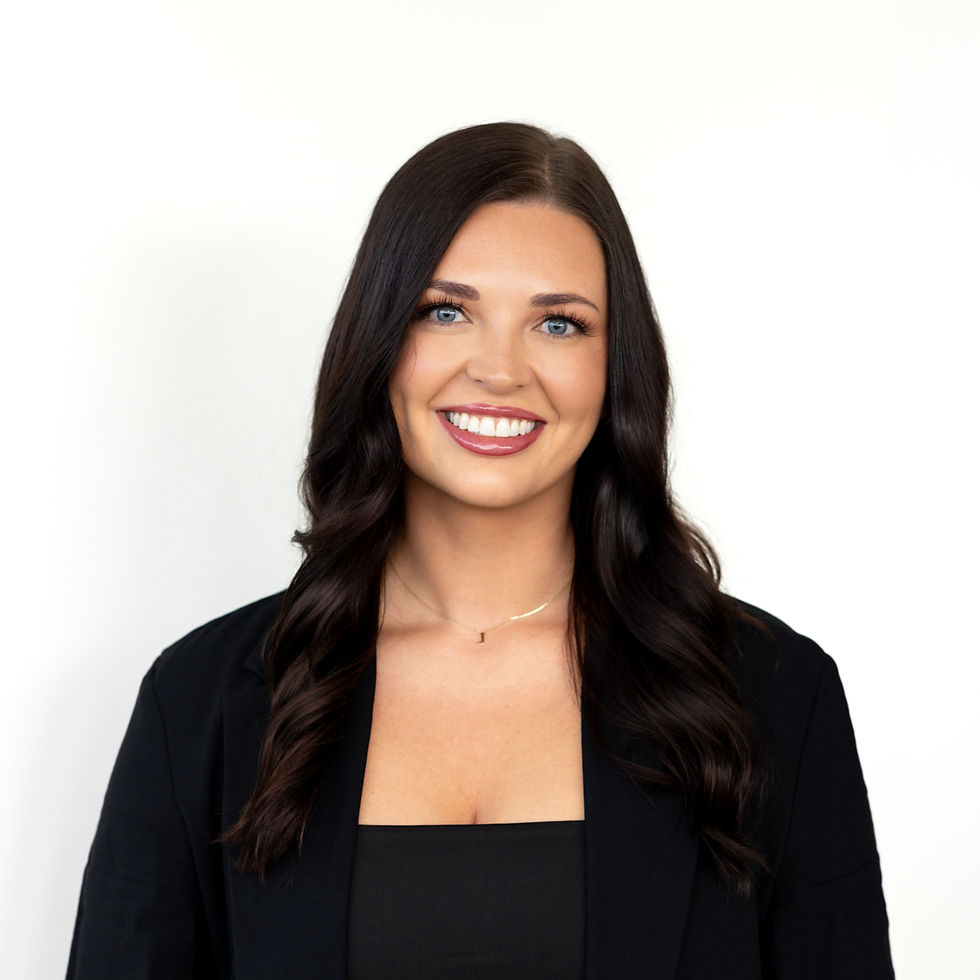 Smiling woman with long dark hair in a black blazer and top, and blue jeans, stands against a plain white background.