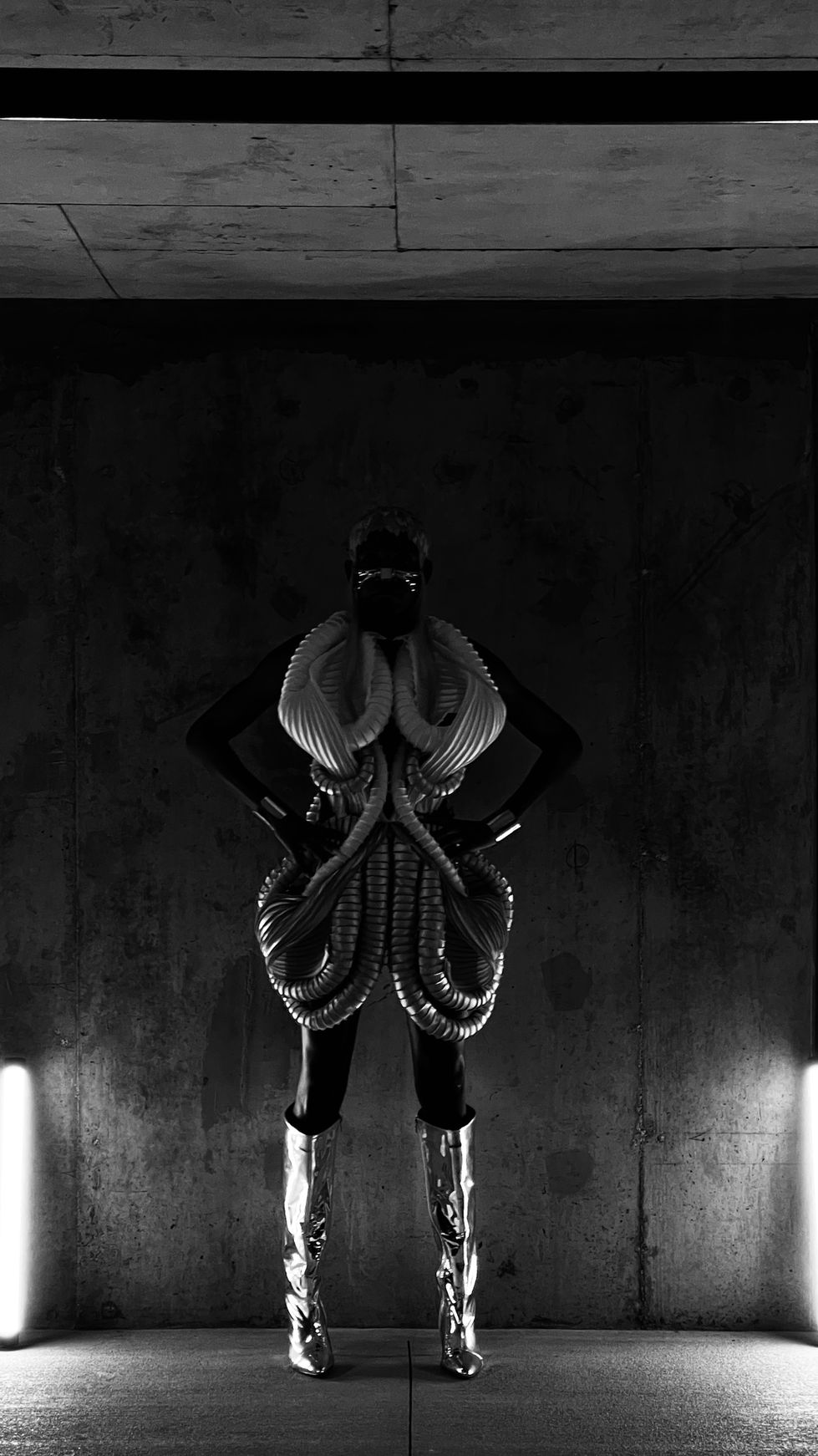 A black and white image, i am in a fabricated dress made of coils and tubes, i looks like it is for an alien. i have shiny silver high heeled boots on, and my hair piece is a mosaic of pastel orange, pink, yellow, and peach. the hair piece looks like paper almost on my head with two long strands of multicolored hair on ether side just in front  of my ears.