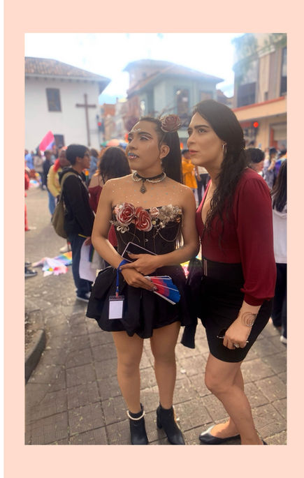 This photo shows two young women standing closely together at a crowded outdoor event. The woman on the left, Emily Amanda Amor, has long dark hair half-up and is wearing a black strapless dress with floral and decorative details, holding fans and her phone. The woman on the right, PJ DiPietro, has long wavy hair and is dressed in a red top with a black skirt, holding sunglasses and some jewelry. Both women appear engaged and observant, with a lively street scene in the background.