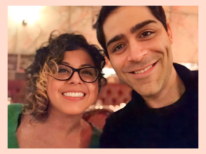 This image features two individuals smiling together in a warmly lit indoor setting. The person on the left has curly hair, glasses, and is wearing a green top, while the person on the right has short dark hair and is dressed in black.