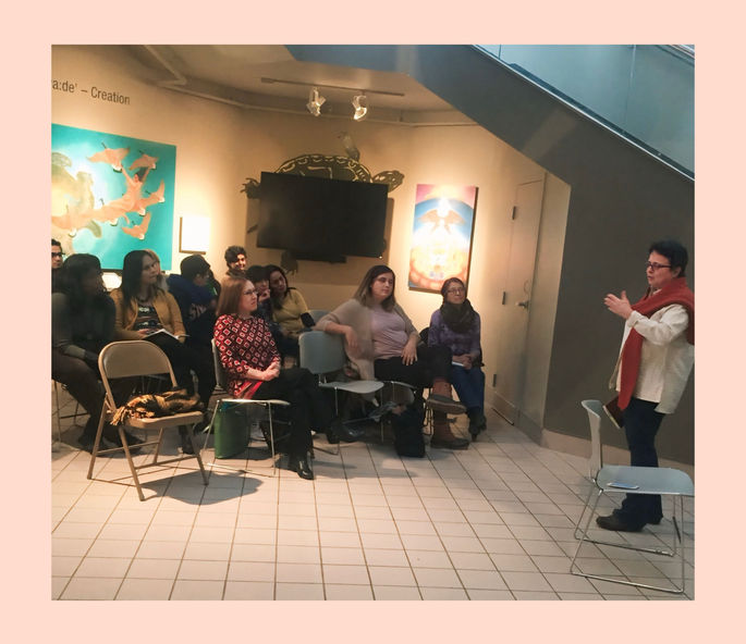 This image shows Maestra Moraga standing in front of an audience, speaking and gesturing with her hands. The audience, consisting of several people, is seated and listening attentively in a cozy, art-filled space.