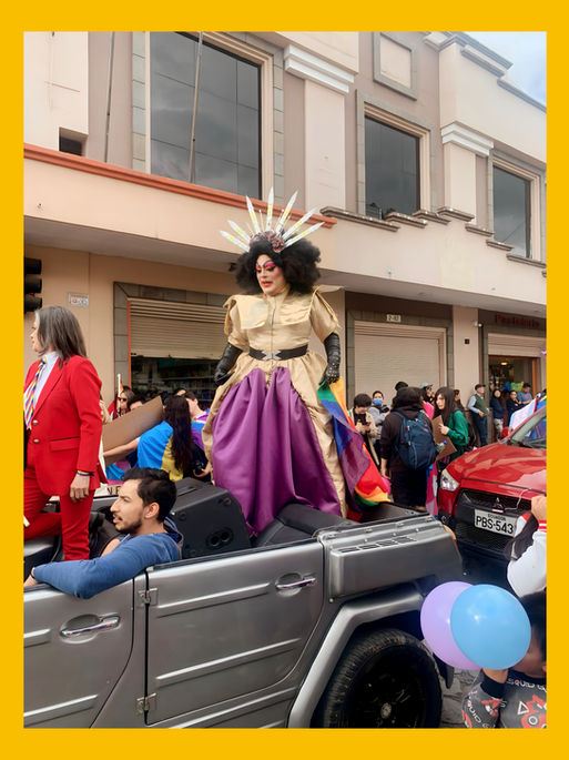 A person in a dramatic costume with a large headdress stands on a silver vehicle during a parade, surrounded by colorful decorations and spectators. The individual wears a gold and purple outfit, showcasing vibrant colors, while others in the background are dressed in festive attire, contributing to the lively atmosphere.