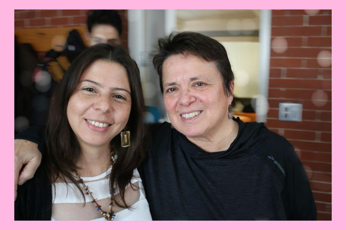 This photo shows two smiling women standing close together indoors, with their arms around each other. The woman on the left, Dr. Arango-Vargas, has long dark hair and freckles, wearing large earrings and a necklace. The woman on the right, Maestra Moraga, has short dark hair and is dressed in a black top.