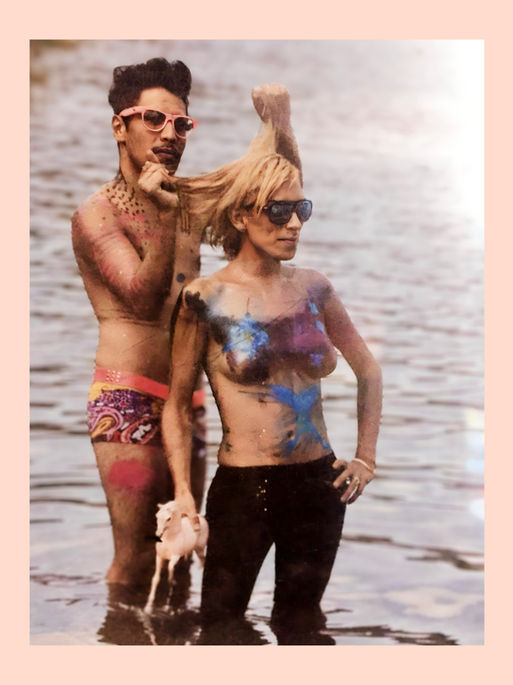 This image depicts two individuals standing in the water, with colorful paint or body art on their upper bodies. The background shows a body of water, and the figures are wearing sunglasses, striking confident poses.