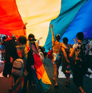 A group of people in party attire walk under a huge rainbow flag, their arms raised.