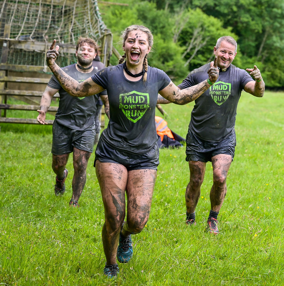 Three muddy runners in "Mud Monsters Run" shirts smile and gesture in a grassy field with trees and a net obstacle in the background.