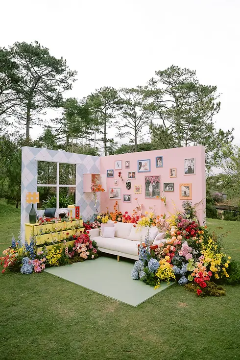 Sân khấu lễ vows đặc biệt ngoài trời với backdrop màu pastel, sofa trung tâm và hoa trang trí rực rỡ trong khu vườn.