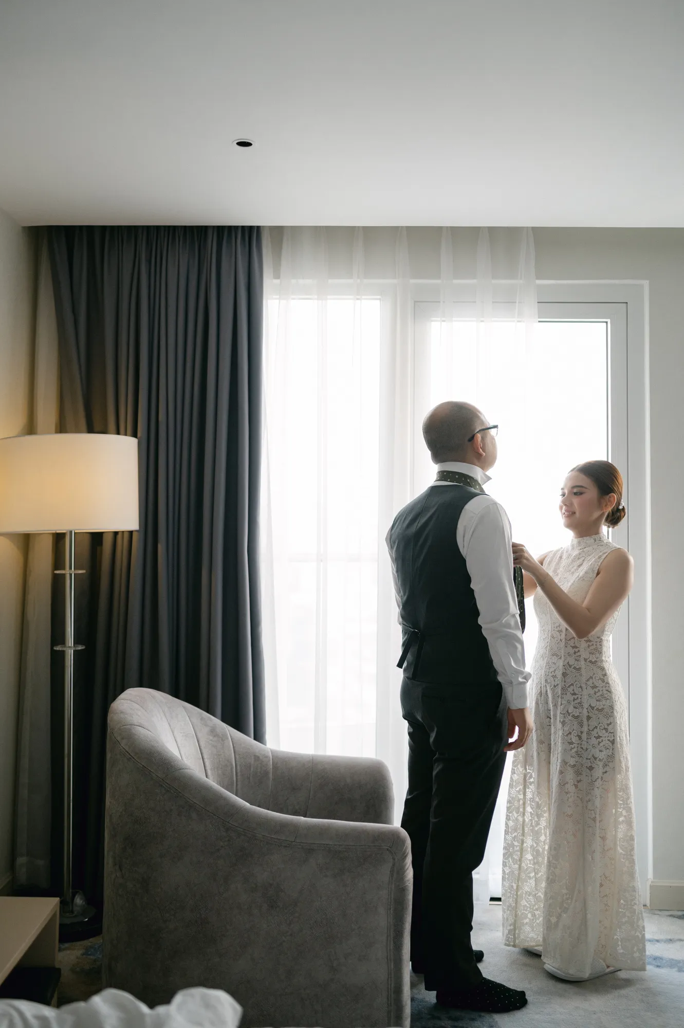 Bride helping the groom adjust his tie in a softly lit hotel room before the wedding ceremony.