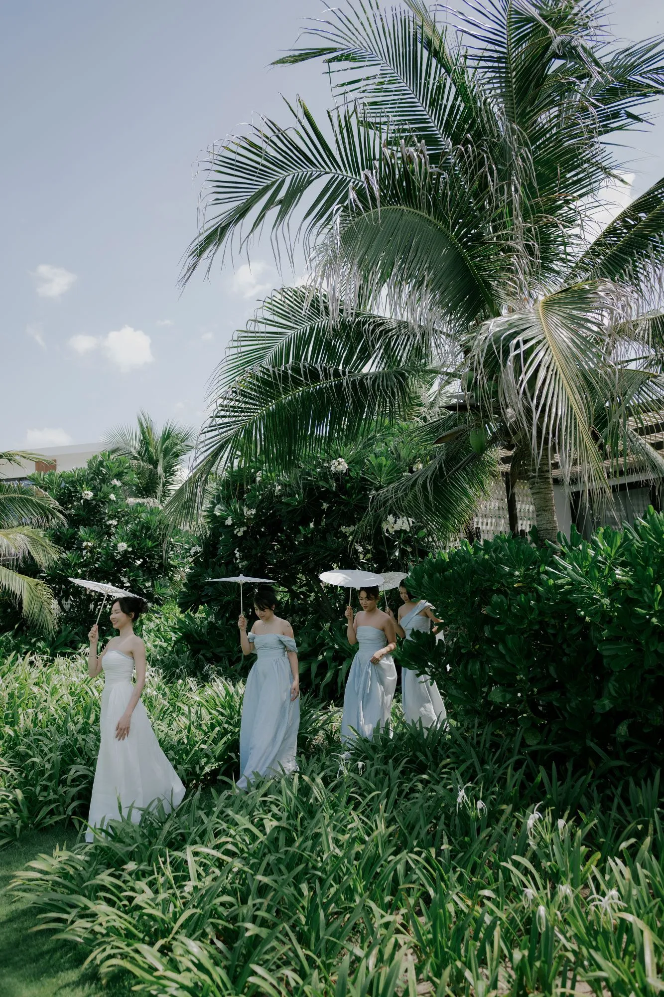 Bridesmaids in soft pastel dresses walk through a lush tropical garden holding white parasols.
