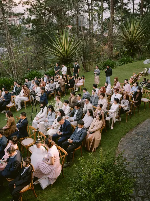 Toàn cảnh khách mời ngồi dự lễ cưới ngoài trời trên bãi cỏ, bố cục nhìn cao cho thấy không gian trang trọng và thoáng đãng.