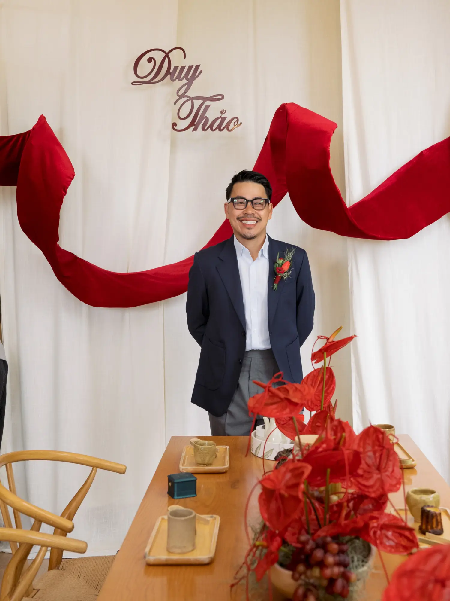 Chú rể mỉm cười rạng rỡ trong không gian lễ gia tiên, phía sau là phông nền “Duy & Thảo” với dải lụa đỏ tượng trưng cho tình yêu và may mắn.