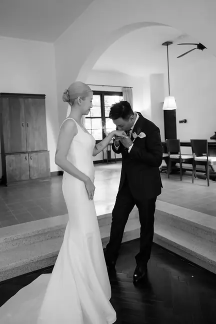 Chú rể cúi hôn tay cô dâu trong không gian tối giản, khung hình đen trắng làm nổi bật sự trang trọng và dịu dàng.