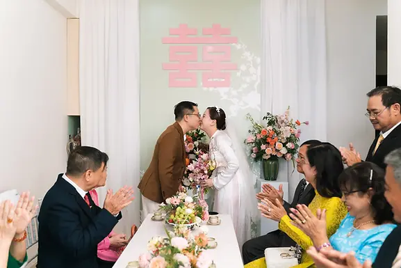Bride and groom sharing a kiss during the tea ceremony while family members applaud.