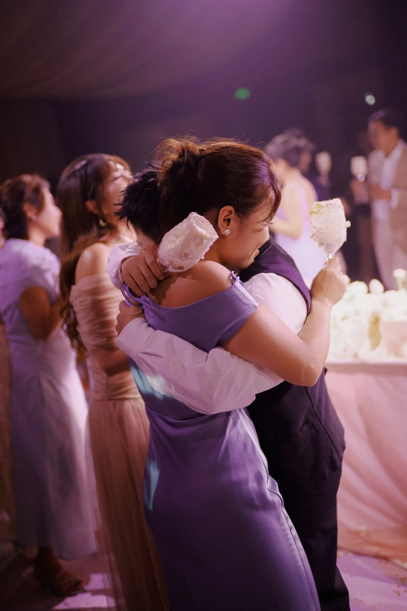 Chú rể và khách mời ôm nhau đầy xúc động trong không gian tiệc cưới lãng mạn dưới ánh đèn tím.